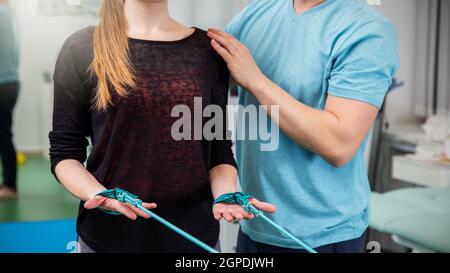 Young sporty woman wearing black top and performs exercises for the muscles of the hands, workout with resistance bands with assistance of physical th Stock Photo