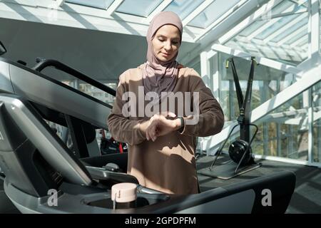 Horizontal medium portrait of modern young adult Muslim woman wearing hijab standing on treadmill in gym checking something on her activity tracker Stock Photo