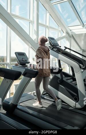 Vertical long shot of unrecognizable young woman wearing hijab running on treadmill exercise machine in modern gym Stock Photo
