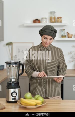 Vertical medium portrait of stylish young Muslim woman wearing turban standing in kitchen using digital tablet Stock Photo