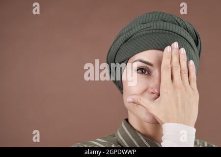 Horizontal close-up studio portrait of attractive Muslim woman wearing stylish shirt and turban covering half of face with hand looking at camera, brown background Stock Photo