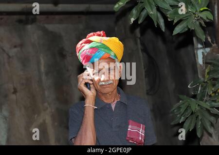 Close-up of Indian-origin farmer standing in field talking on mobile phone and smiling wearing colorful turban according to Rajasthani tradition and H Stock Photo
