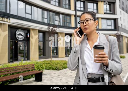 Young pretty female manager in suit having drink and phoning against architecture Stock Photo