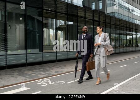 Two happy young intercultural colleagues walking along modern architecture and discussing latest news Stock Photo