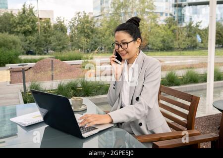 Young successful businesswoman using mobile phone and laptop during work in outdoor cafe Stock Photo