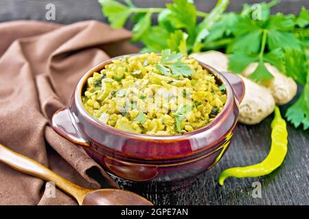 Indian national dish kichari made of mung bean, rice, celery, spinach, hot pepper and spices in a bowl on a towel, ginger and spoon on dark wooden boa Stock Photo