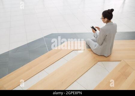 Young restful businesswoman in grey suit listening to music while sitting on large steps Stock Photo