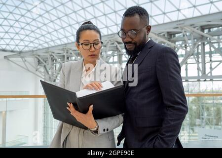 Young Asian businesswoman in formalwear showing financial papers or contract to African colleague Stock Photo