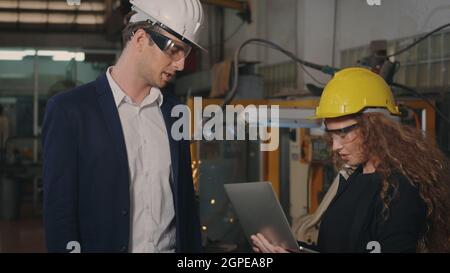 Two heavy industry business engineers in hardhats discuss information on laptop computer while standing indoors welding manufacturing industrial Stock Photo