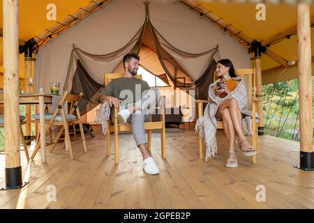 Young relaxed couple having talk while sitting in armchairs on patio of glamping tent Stock Photo