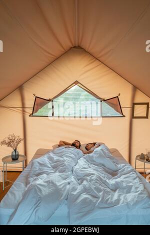 Young couple sleeping under white blanket on double bed in bedroom of glamping tent Stock Photo
