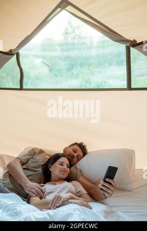 Young restful couple with smartphone making video call while relaxing in bed inside glamping house Stock Photo