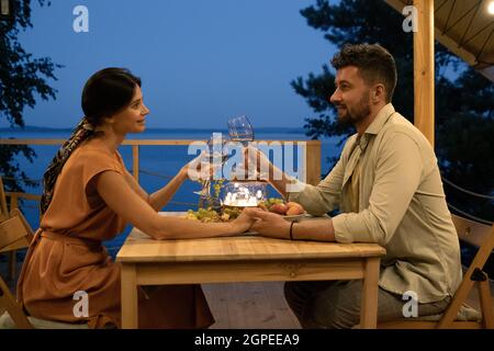Happy couple with glasses of wine toasting by served table while enjoying evening by seaside Stock Photo