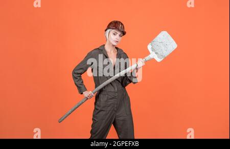 woman hard worker in protective helmet and boilersuit hold shovel on orange background, labour day Stock Photo