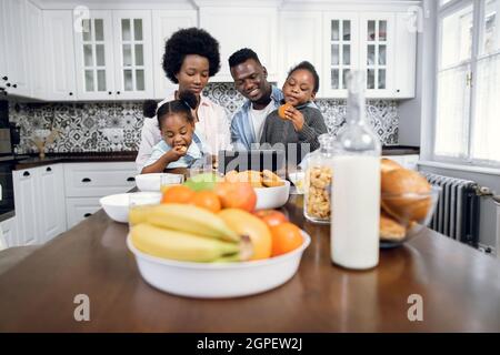 Two pretty sisters sitting on parent's knees while watching cartoon on bright kitchen. African american family using digital tablet during tasty breakfast at home. Stock Photo
