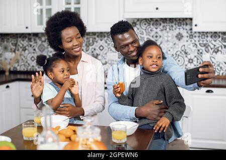 Cheerful parents sitting on bright kitchen with their two cute daughters and taking selfie on cell phone. Happy african american family spending morning time at home with pleasure and fun. Stock Photo
