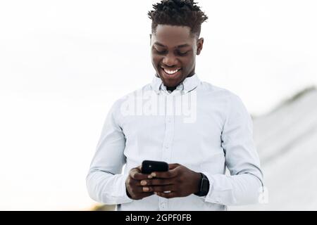 Happy fashionable curly African man, in white shirt, using mobile phone, outdoors on the street, business concept, work, gadgets Stock Photo