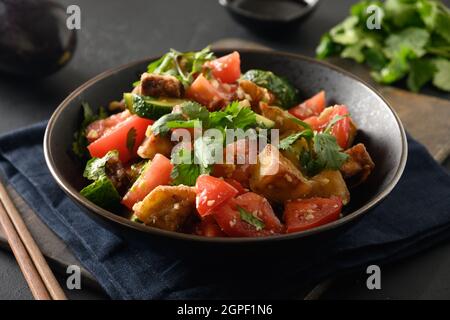 Chinese crispy spicy eggplant with cucumber, garlic, tomatoes, fresh cilantro leaves in oyster sauce and black vinegar on black background. Close up. Stock Photo