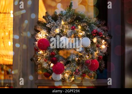 Christmas wreath made of fir branches with round balls of red, white and golden color and luminous garlands on the door. Decorating the door with a be