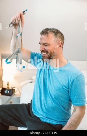 Mature happy man with chronic breathing issues considers using CPAP machine sitting on the bed in bedroom . Healthcare, CPAP, snoring concept Stock Photo