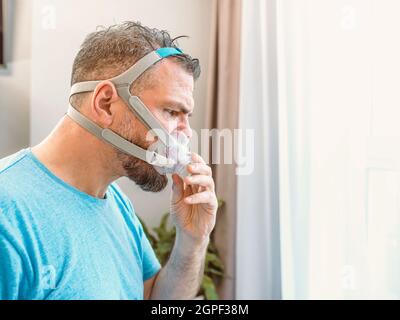 Mature happy man with chronic breathing issues considers using CPAP machine sitting on the bed in bedroom . Healthcare, CPAP, snoring concept Stock Photo