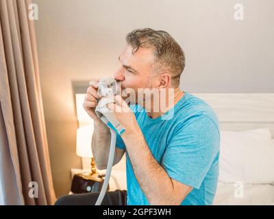 Mature happy man with chronic breathing issues considers using CPAP machine sitting on the bed in bedroom . Healthcare, CPAP, snoring concept Stock Photo