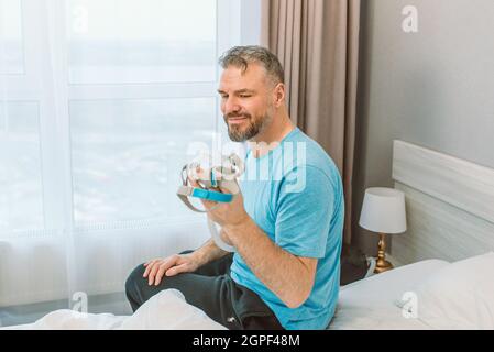 Mature happy man with chronic breathing issues considers using CPAP machine sitting on the bed in bedroom . Healthcare, CPAP, snoring concept Stock Photo