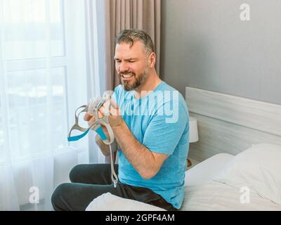 Mature happy man with chronic breathing issues considers using CPAP machine sitting on the bed in bedroom . Healthcare, CPAP, snoring concept Stock Photo