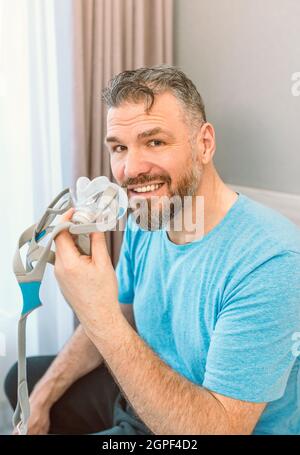 Mature happy man with chronic breathing issues considers using CPAP machine sitting on the bed in bedroom . Healthcare, CPAP, snoring concept Stock Photo