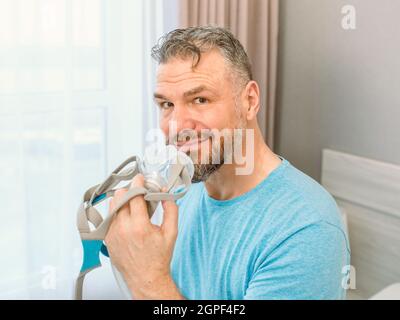 Mature happy man with chronic breathing issues considers using CPAP machine sitting on the bed in bedroom . Healthcare, CPAP, snoring concept Stock Photo