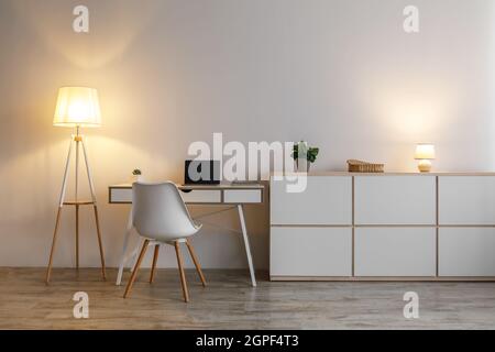 Cozy modern workplace in evening with table, computer, armchair and glowing lamp, on gray wall background Stock Photo