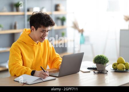 Smiling asian man working on laptop and writing Stock Photo