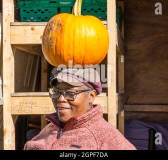 Senior woman selling pumpkins balancing a pumpkin on her head at her market stall close up facing camera. Stock Photo