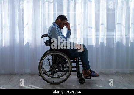 Side view of paraplegic black man feeling desperate and lonely, having depression, sitting in wheelchair near window Stock Photo