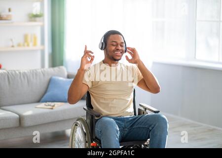 Cheery disabled black guy in wheelchair wearing headphones, listening to music with closed eyes at home Stock Photo