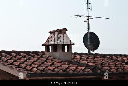 A stone chimney covered with brick tiles next to a satellite antenna on the roof of a building (Umbria, Italy, Europe) Stock Photo