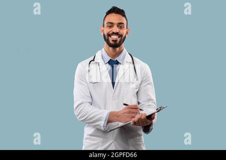 Happy Middle-Eastern Doctor Man Taking Notes Standing Over Blue Background Stock Photo