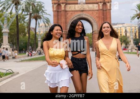 Group of delighted young multiracial ladies in stylish summer outfits walking together on city square with triumphal arch in background