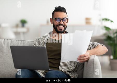 Young Arab guy making video call, reading documents, using laptop, working from home office Stock Photo