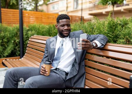 Smiling african american manager holding takeaway coffee and using smartphone while sitting on bench outdoors Stock Photo