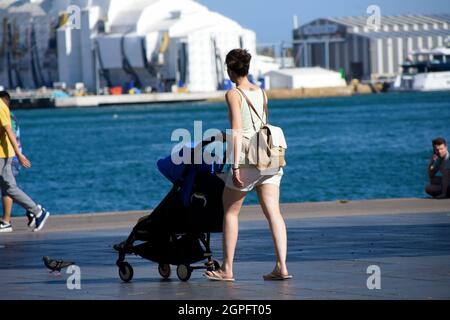 Mother pushing a stroller with her baby inside. She is walking in a promenade near the sea. Stock Photo