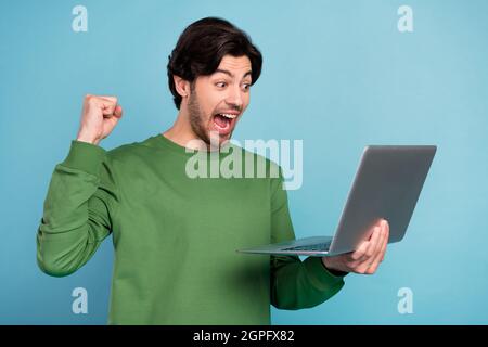 Portrait of young excited crazy cheerful guy raise fist in triumph victory using laptop isolated on blue color backgroung Stock Photo