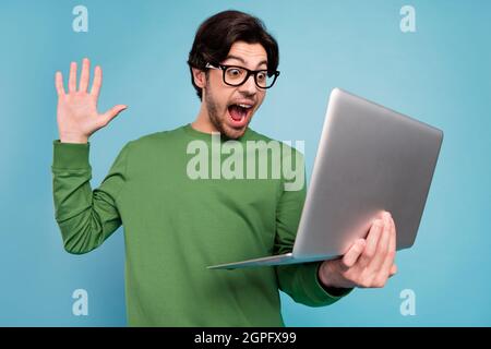 Portrait of young excited crazy cheerful guy in glasses scream in triumph victory using laptop isolated on blue color background Stock Photo