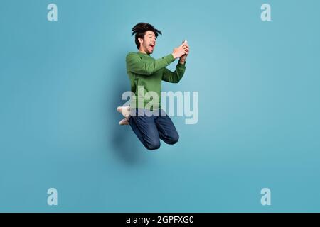Full size profile side photo of crazy excited handsome man jumping using smartphone isolated on blue color background Stock Photo
