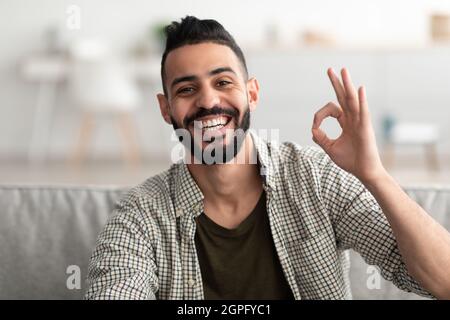 Happy young Arab guy showing okay gesture and smiling at camera, sitting on sofa at home Stock Photo