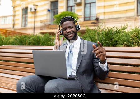 Excited african american businessman in wireless headphones using modern laptop and listening music, sitting outdoors Stock Photo