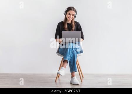 Cheery Caucasian woman in headphones having online video call on laptop, sitting on chair against white wall Stock Photo