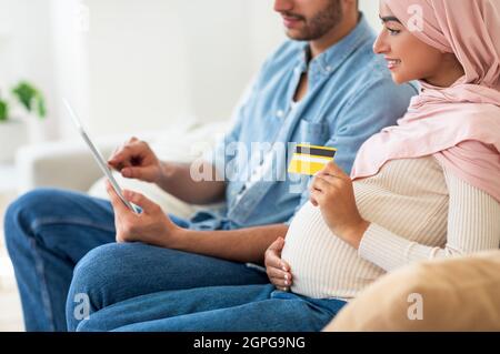 Online shopping for pregnant couple. Muslim man and woman using digital tablet and credit card, buying things for baby Stock Photo