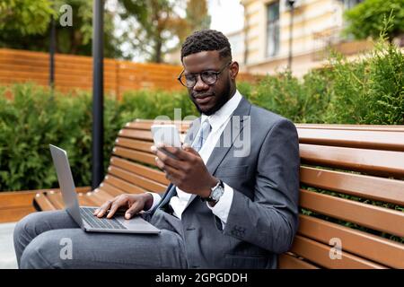 Young african american businessman with laptop sitting on bench outdoors and using smartphone, free space Stock Photo