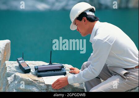 Singapore. Technology. Engineer outdoors using satellite phone with printer. Stock Photo